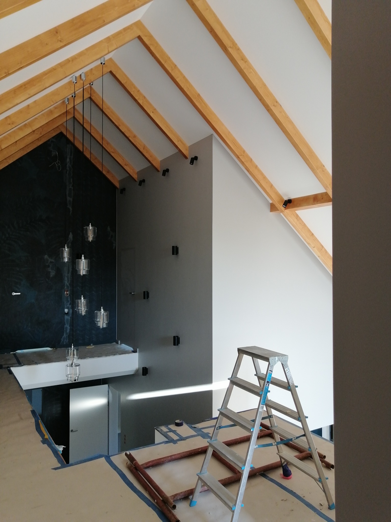Poddasze w trakcie prac wykończeniowych: widoczne drewniane belki stropowe, pomalowane ściany w odcieniach szarości, nowoczesne lampy wiszące oraz aluminiowa drabina rozstawiona na zabezpieczonej...