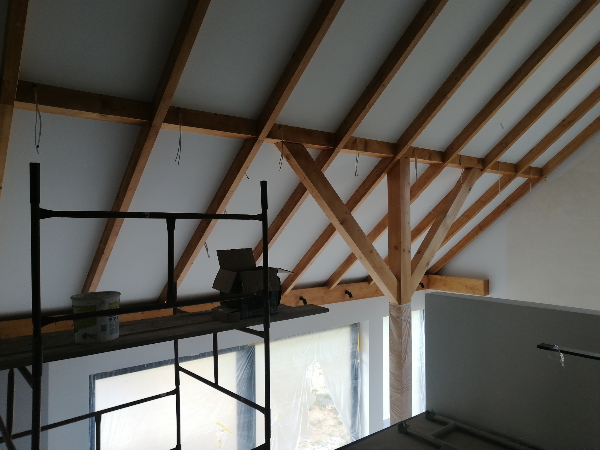 Poddasze w trakcie ocieplania, widoczne drewniane krokwie, biała wełna mineralna między nimi, rusztowanie z wiadrem farby i kartonami, okno zabezpieczone folią.