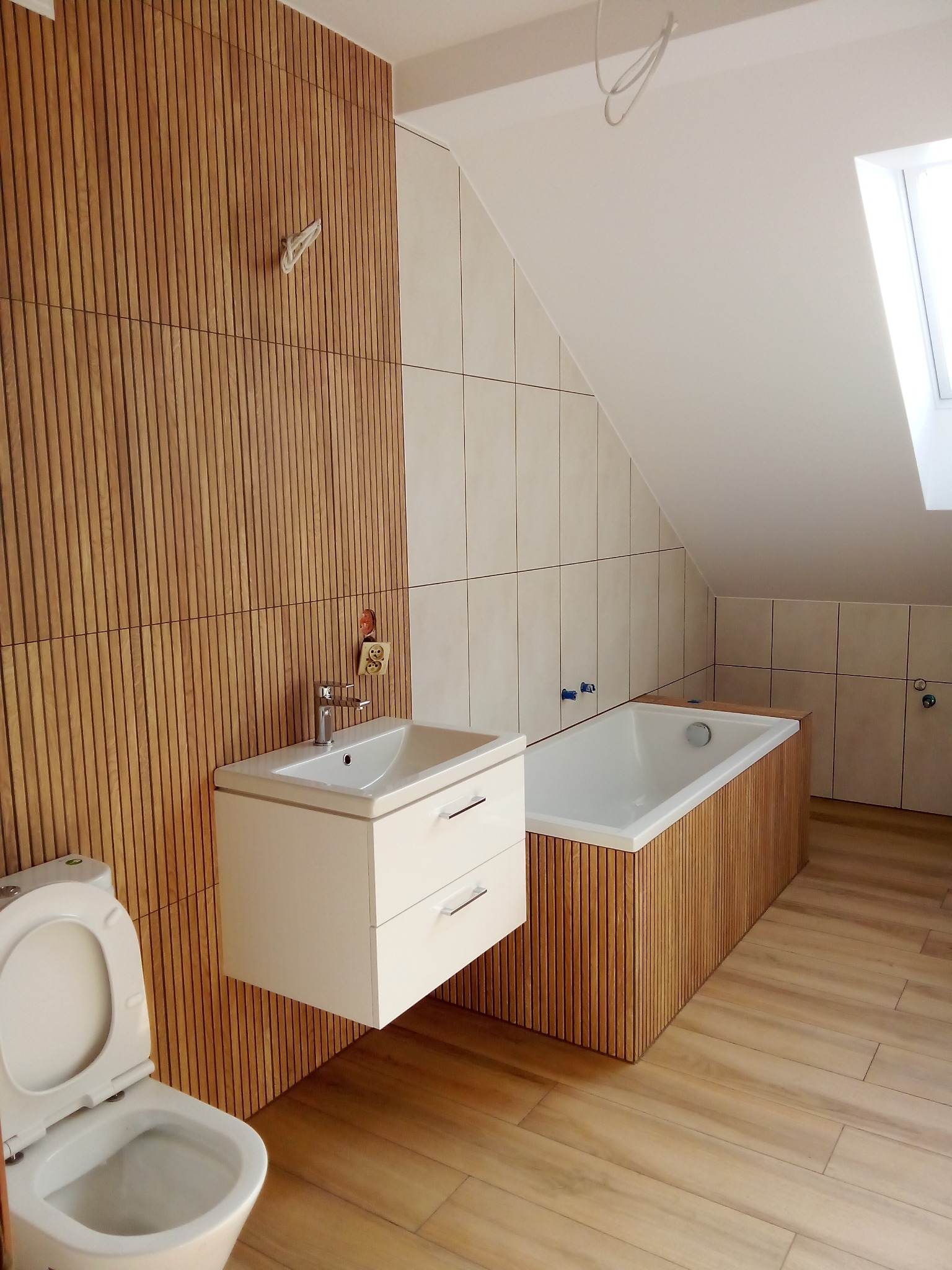 Ukończona łazienka na poddaszu z wanną obudowaną drewnianymi lamelami, wiszącą szafką z umywalką i toaletą, drewnopodobne płytki na podłodze i ścianach.