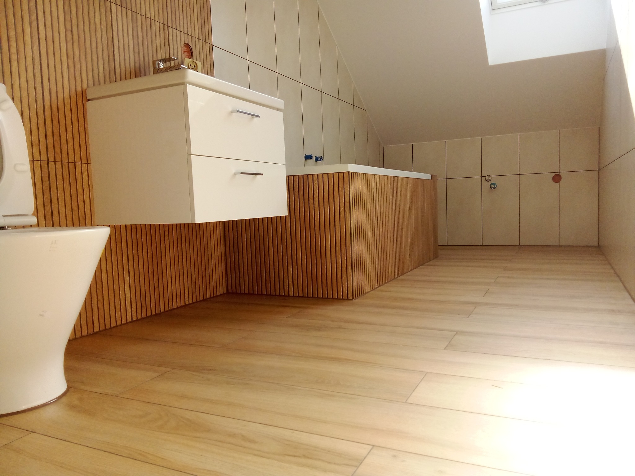 Nowoczesna łazienka na poddaszu z drewnopodobnymi płytkami na podłodze i ścianach, biała wisząca szafka z umywalką i toaleta.