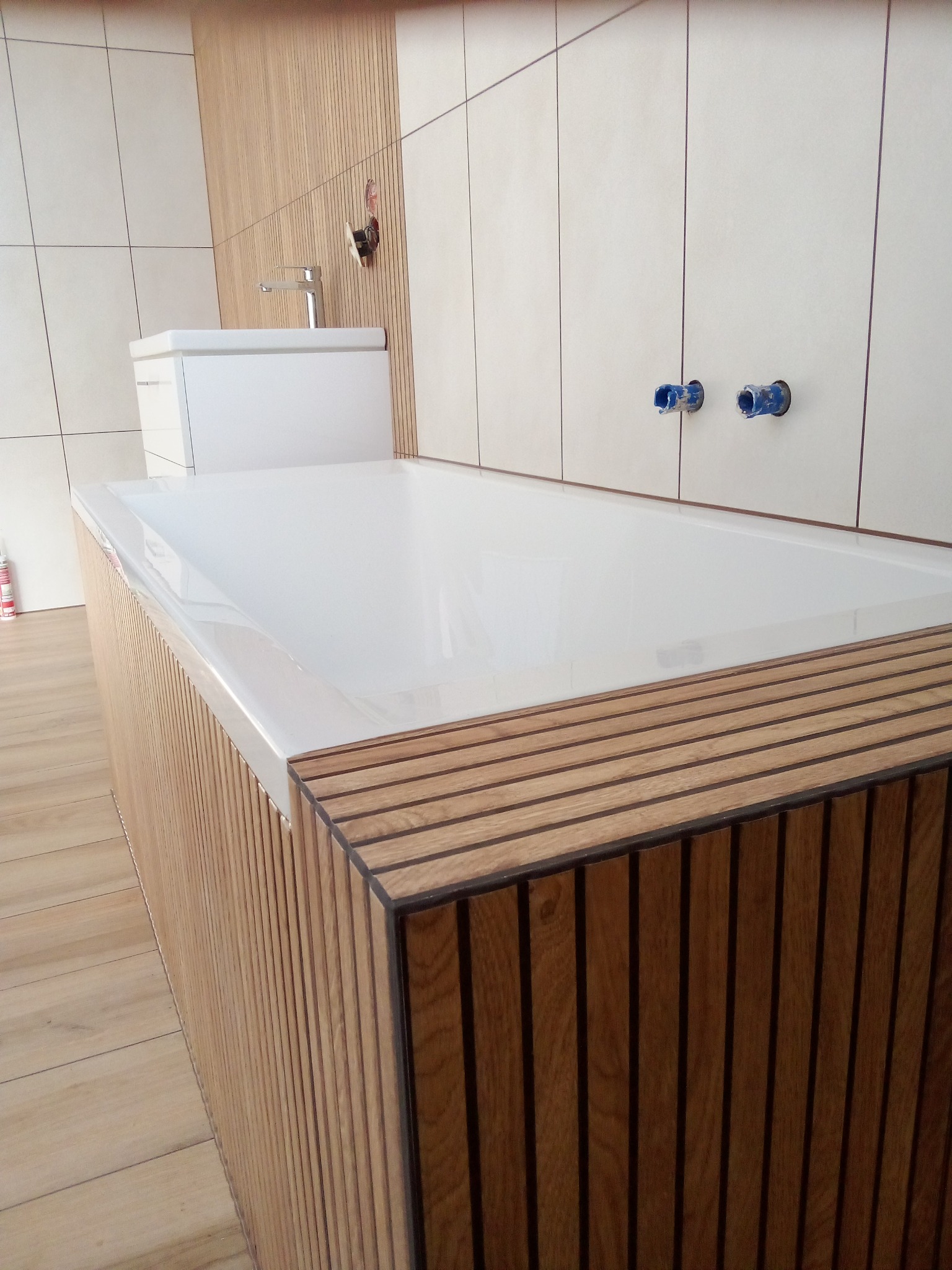 Nowoczesna łazienka z wanną obudowaną drewnianymi lamelami, biała umywalka z chromowaną baterią, i płytki w odcieniach beżu, widoczne niezaślepione przyłącza wodne na ścianie.