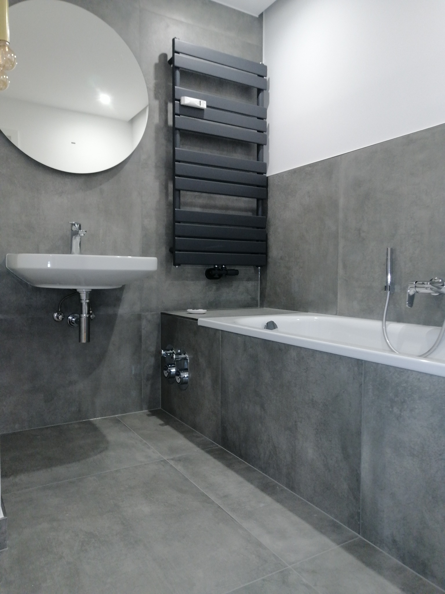 Nowoczesna łazienka z szarymi płytkami, okrągłym lustrem nad białą umywalką i czarnym grzejnikiem drabinkowym, minimalistyczny design.