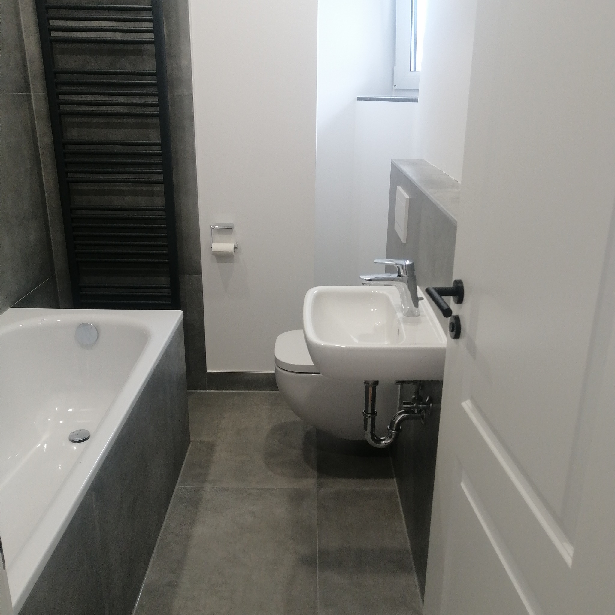 Nowoczesna, minimalistyczna łazienka w szaro-białej kolorystyce z wiszącą umywalką, toaletą, grzejnikiem drabinkowym i wanną, widoczna z perspektywy otwartych drzwi.