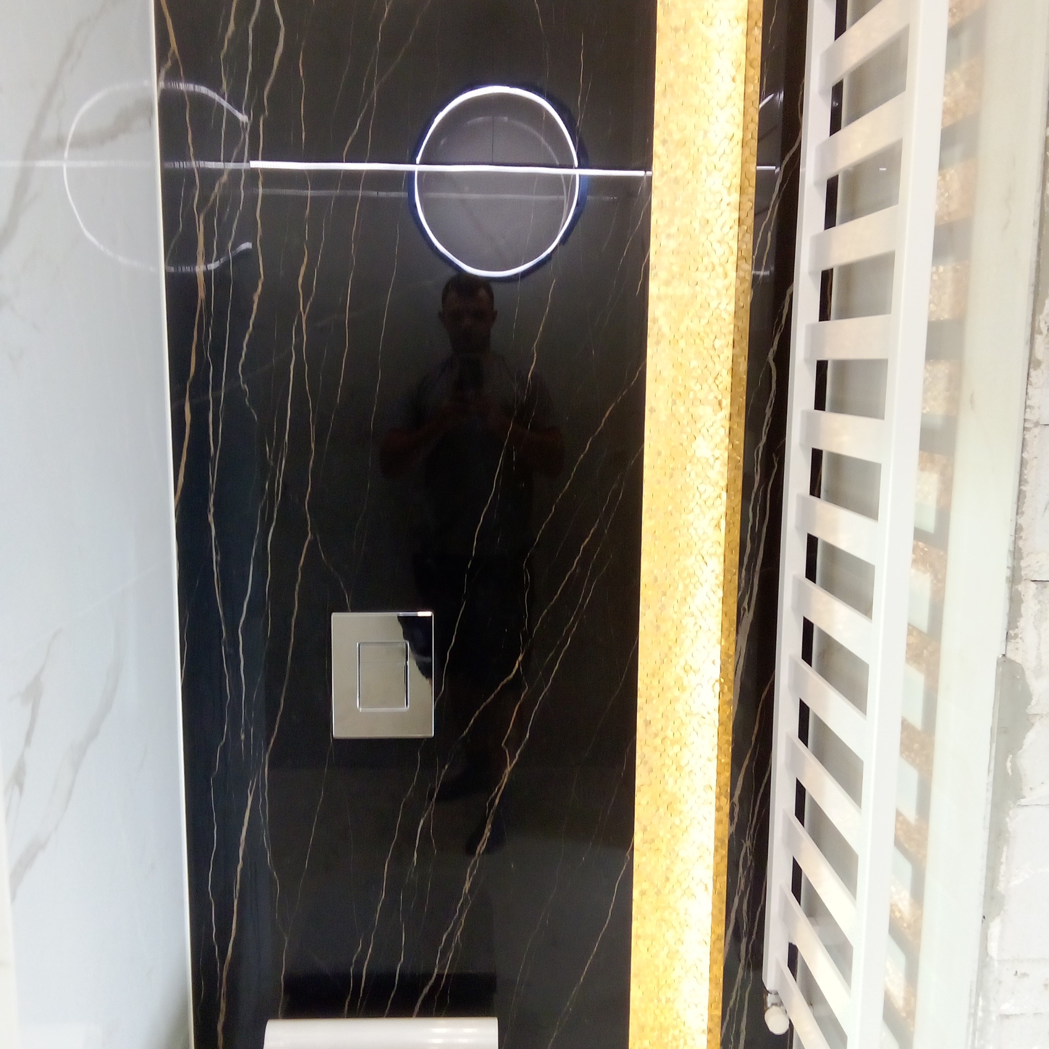 Część ściany w łazience wykończona czarnymi płytkami z złotymi żyłami, podświetlany okrągły przycisk spłuczki, fragment grzejnika drabinkowego i złota mozaika.