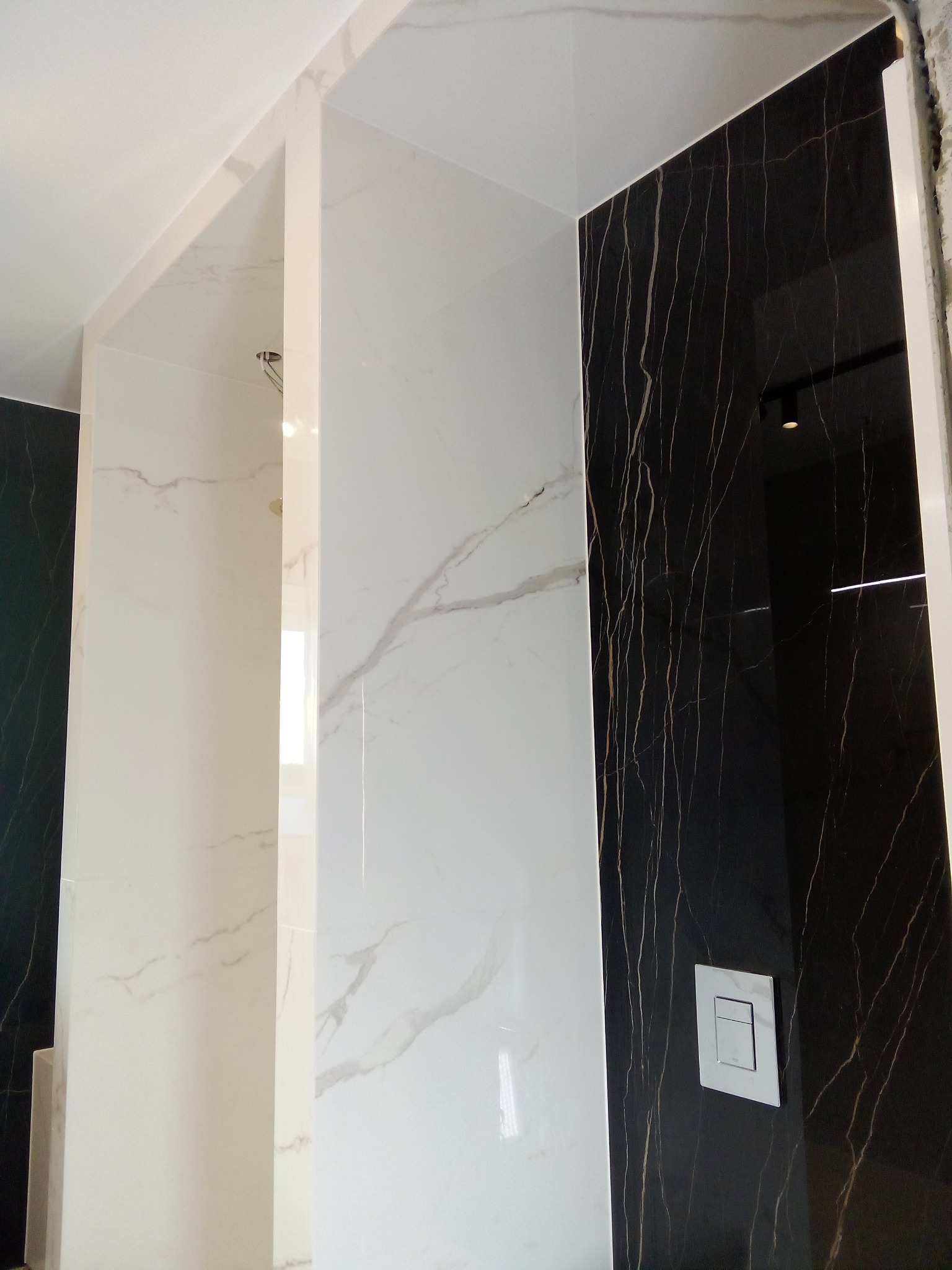 Wykończona łazienka z czarno-białymi płytkami imitującymi marmur, widoczny przycisk spłuczki na czarnej ścianie i fragment sufitu z oświetleniem.