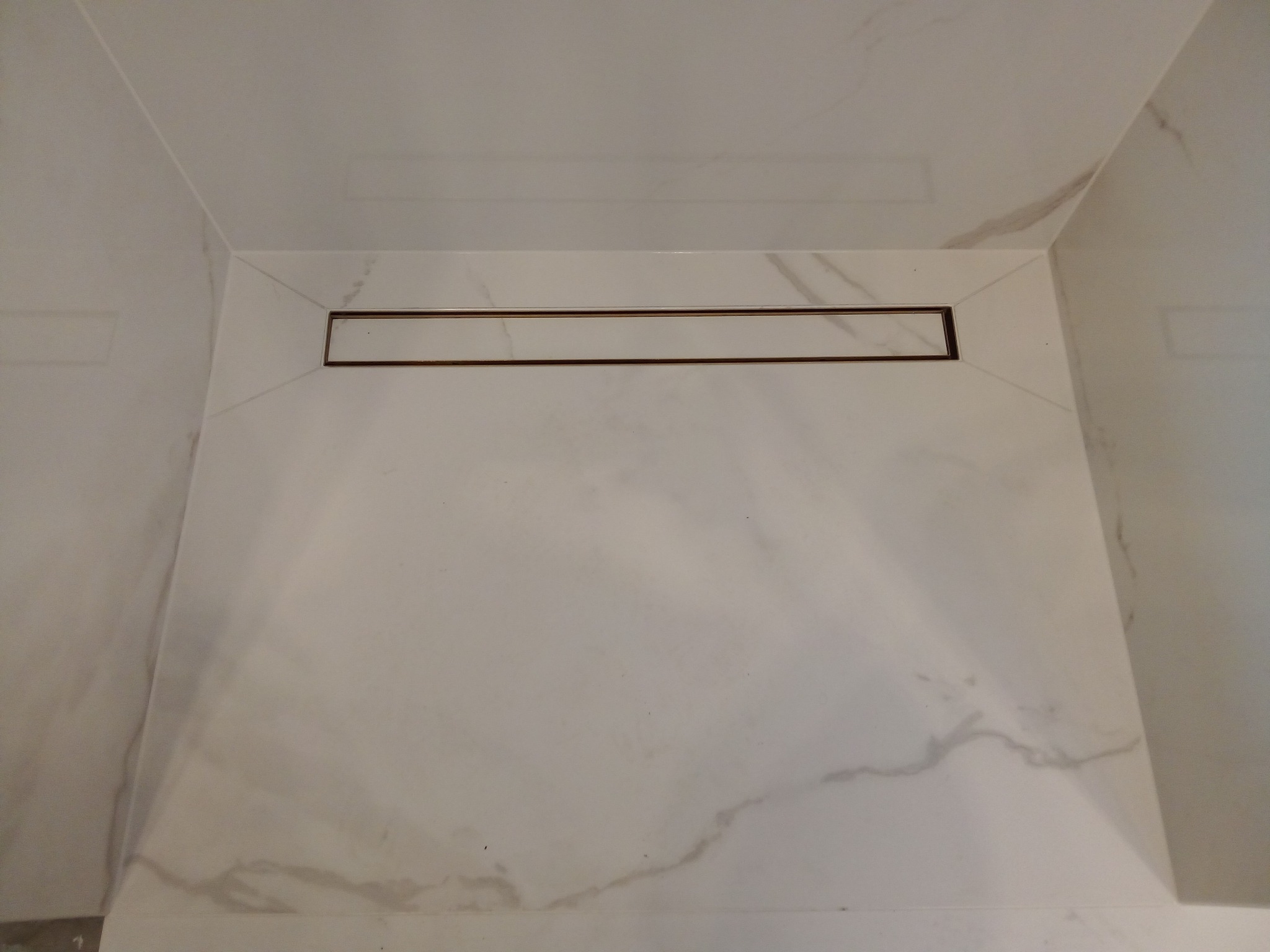 Białe marmurowe płytki w kabinie prysznicowej z odpływem liniowym z brązową ramką.