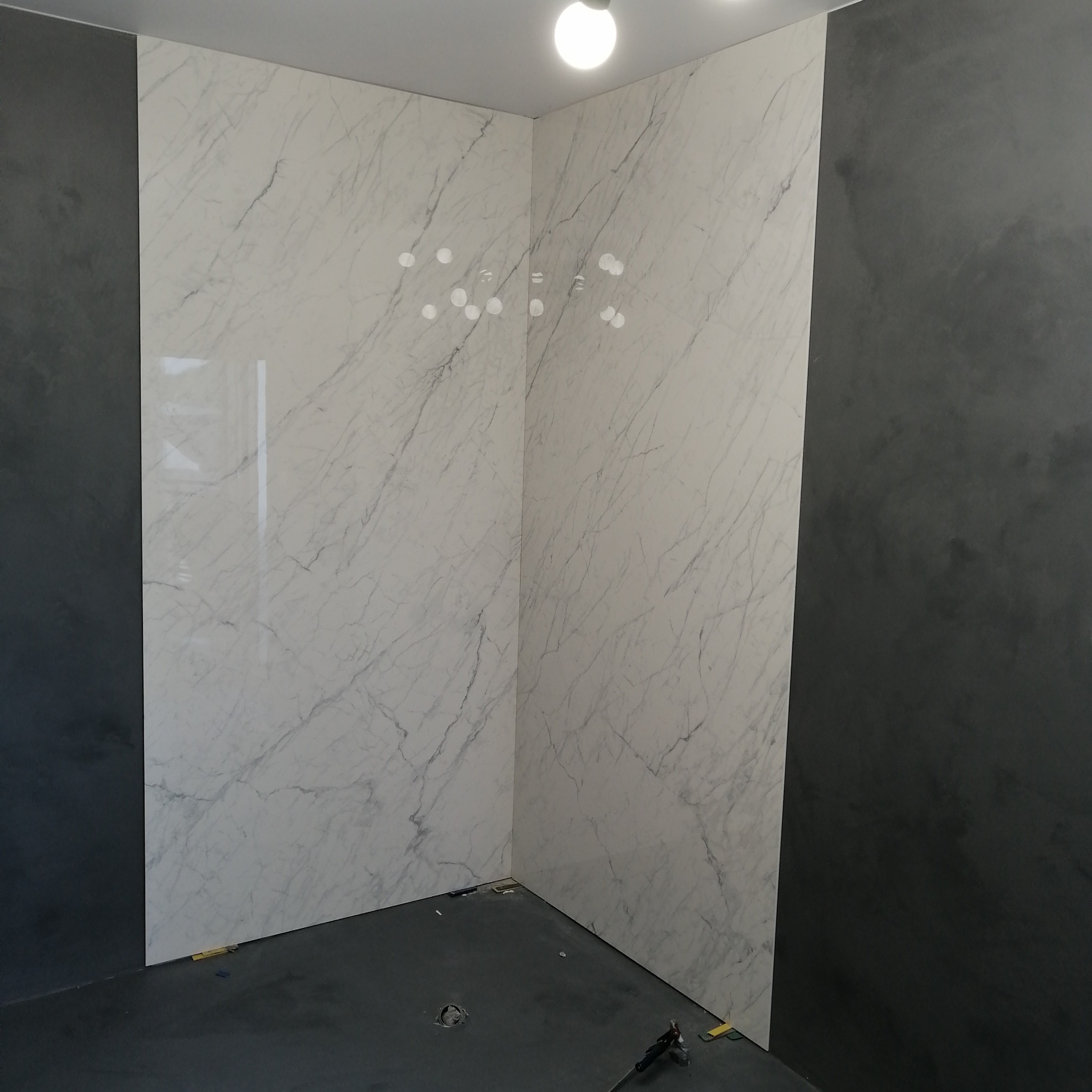 Wykończony narożnik łazienki z płytkami imitującymi biały marmur na ścianach i ciemnoszarą farbą dekoracyjną po bokach, widoczne narzędzia na podłodze.