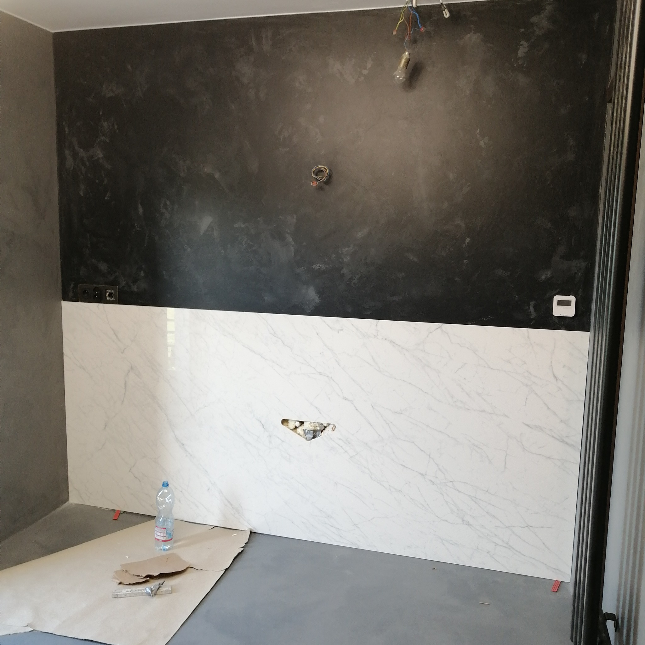 Ściana w trakcie remontu z czarną, teksturowaną górą i marmurowymi płytami na dole, widoczne puszki elektryczne, butelka wody i narzędzia na podłodze zabezpieczonej kartonem.