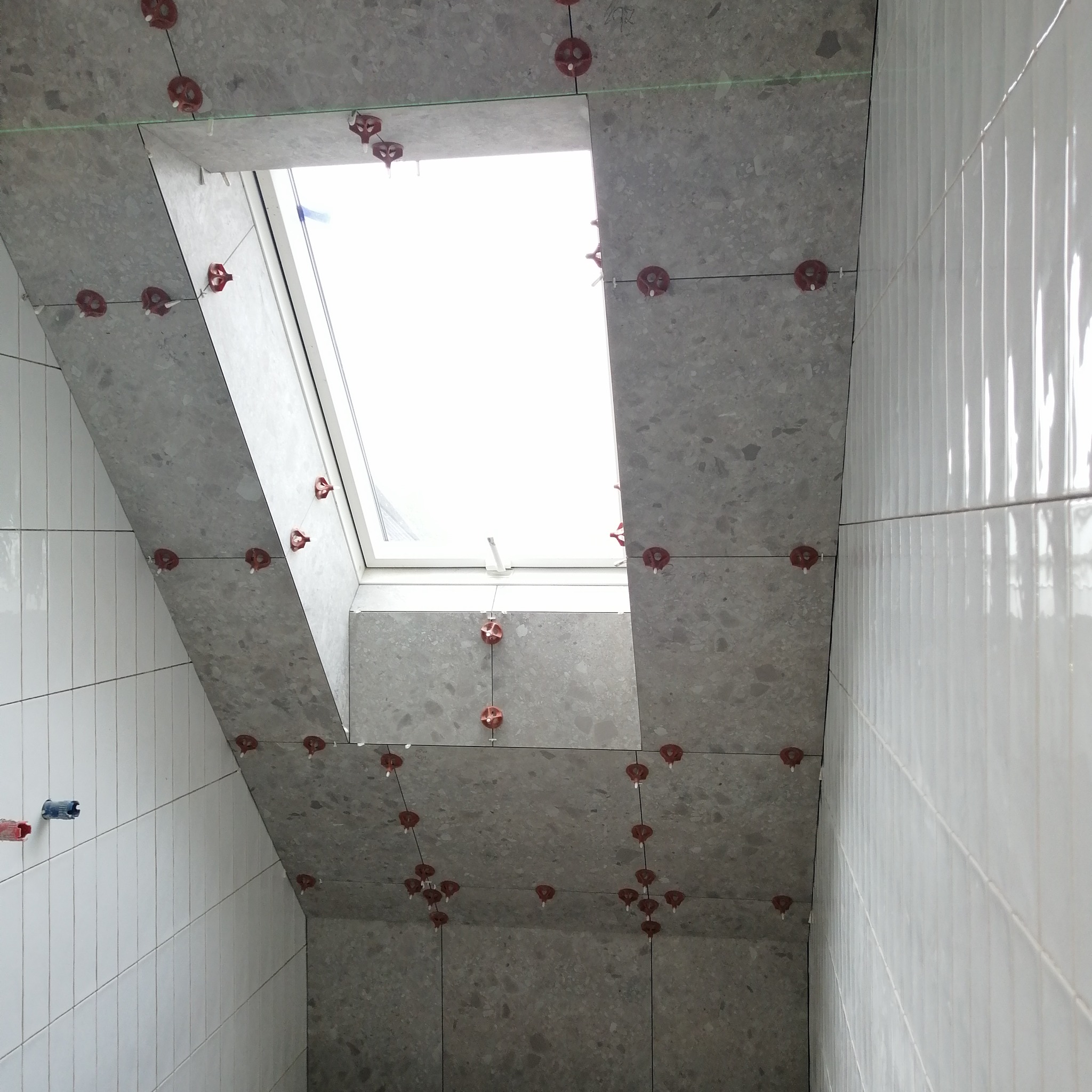 Montaż szarych płytek na poddaszu wokół okna dachowego, widoczne krzyżyki dystansowe i poziomica laserowa.