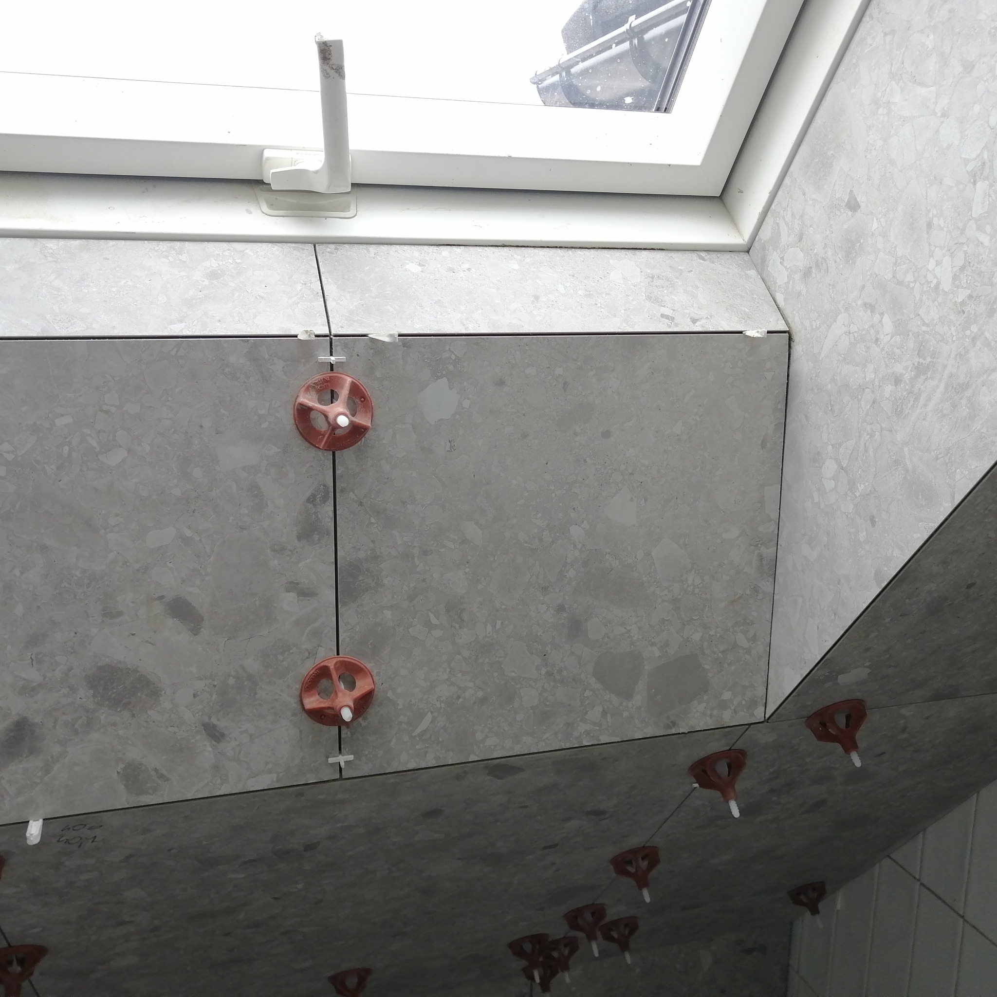 Szare płytki ceramiczne układane pod oknem dachowym, z użyciem pomarańczowych klinów i krzyżyków dystansowych do precyzyjnego poziomowania.