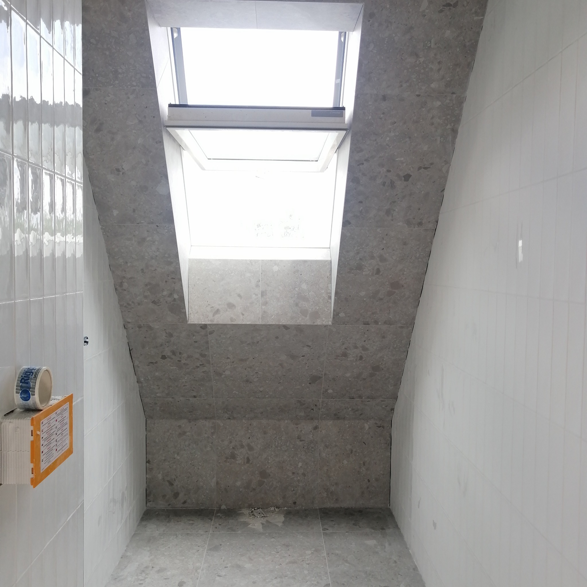 Ściana łazienki z oknem dachowym, wyłożona częściowo białymi, podłużnymi płytkami, a częściowo szarymi płytkami imitującymi lastryko, z widoczną rolką taśmy malarskiej i paczką płytek na ścianie.