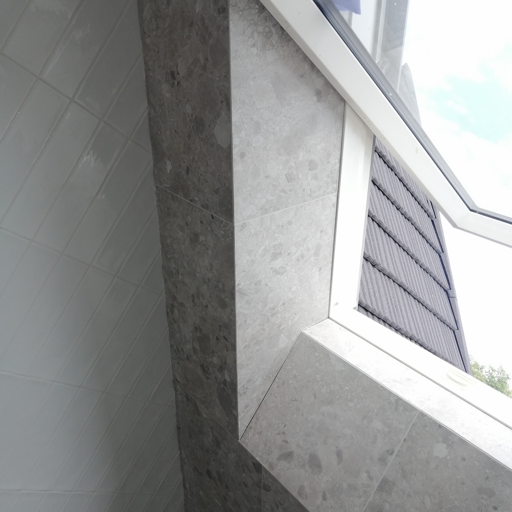 Wykończone obłożenie okna mozaiką z szarego kamienia naturalnego, widoczne białe płytki na ścianie po lewej stronie oraz fragment dachu za oknem.