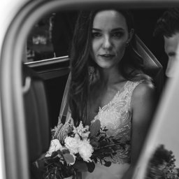 ArtLu Fotografia - Czarno-białe zdjęcie panny młodej w sukni ślubnej z bukietem, siedzącej w samochodzie i patrzącej w obiektyw.