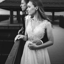 ArtLu Fotografia - Czarno-białe zdjęcie pary młodej stojącej na drewnianym pomoście, z widokiem na pawilon w tle. Panna młoda w koronkowej sukni, pan młody w smokingu z muchą, oboje patrzą w dal.