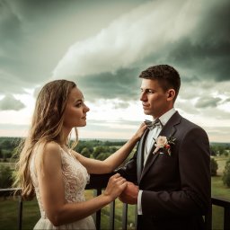 ArtLu Fotografia - Para młoda na balkonie, ona w koronkowej sukni, on w grafitowym smokingu z butonierką, trzymają się za ręce na tle burzowego nieba i zielonego krajobrazu.