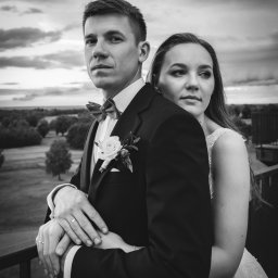 ArtLu Fotografia - Czarno-białe zdjęcie pary młodej na tle pochmurnego nieba i zieleni. Pan młody w ciemnym garniturze z muszką i butonierką, obejmuje pannę młodą w białej sukni. Widoczne obrączki na palcach.