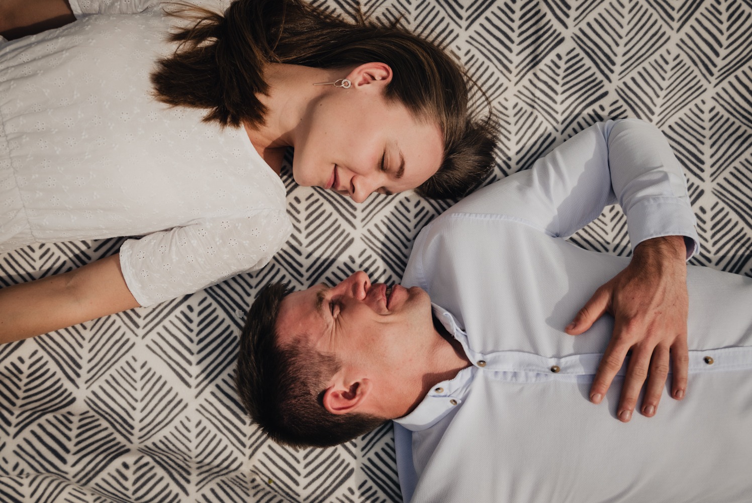 Para leży na wzorzystym kocu, kobieta w białej bluzce z haftem, mężczyzna w jasnoniebieskiej koszuli, oboje z zamkniętymi oczami, delikatny uśmiech na twarzy kobiety.