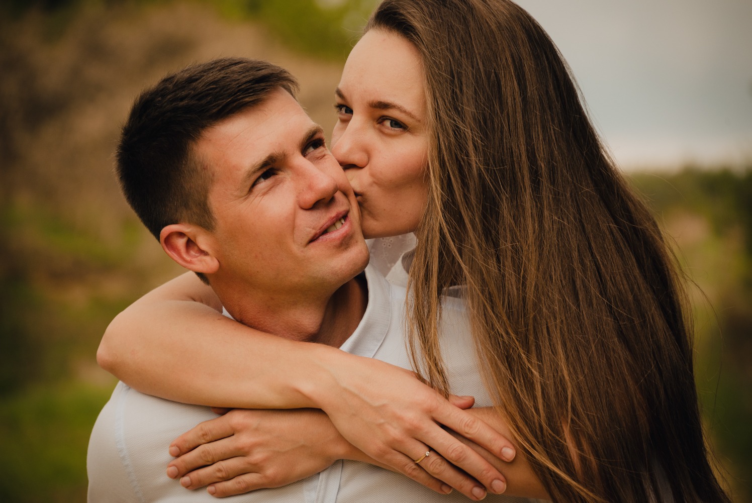 Kobieta całuje mężczyznę w policzek, obejmując go ramionami. Na palcu kobiety widoczny pierścionek zaręczynowy, a w tle zamazane zielone drzewa.