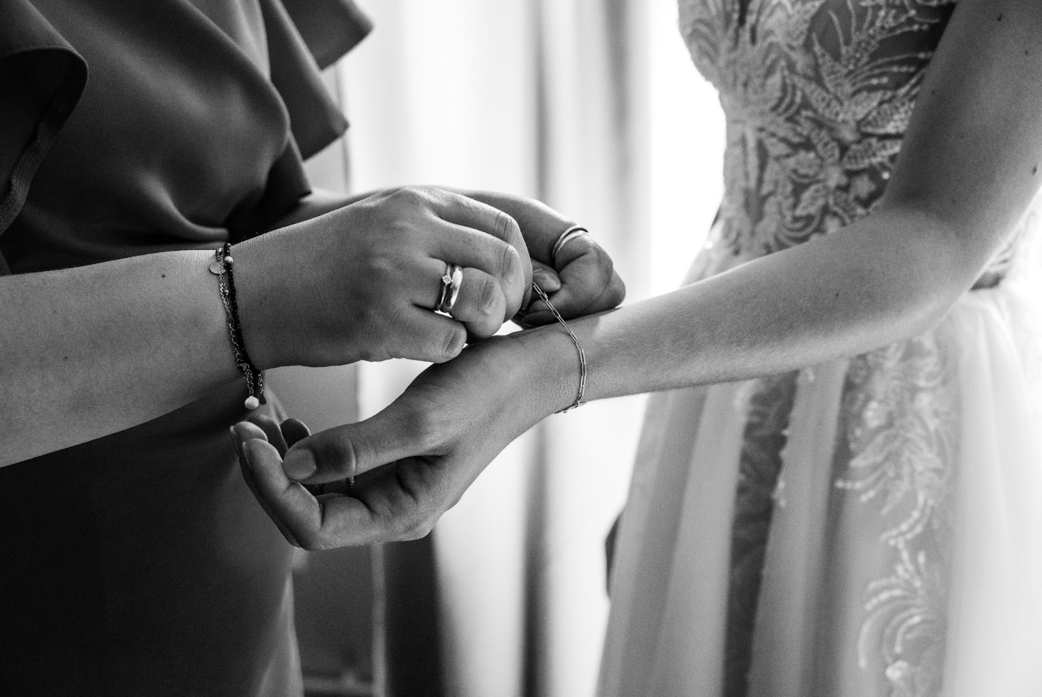 Czarno-białe zdjęcie dłoni zapinających delikatną bransoletkę na nadgarstku panny młodej, która ma na sobie elegancką suknię ślubną z koronkowymi zdobieniami.