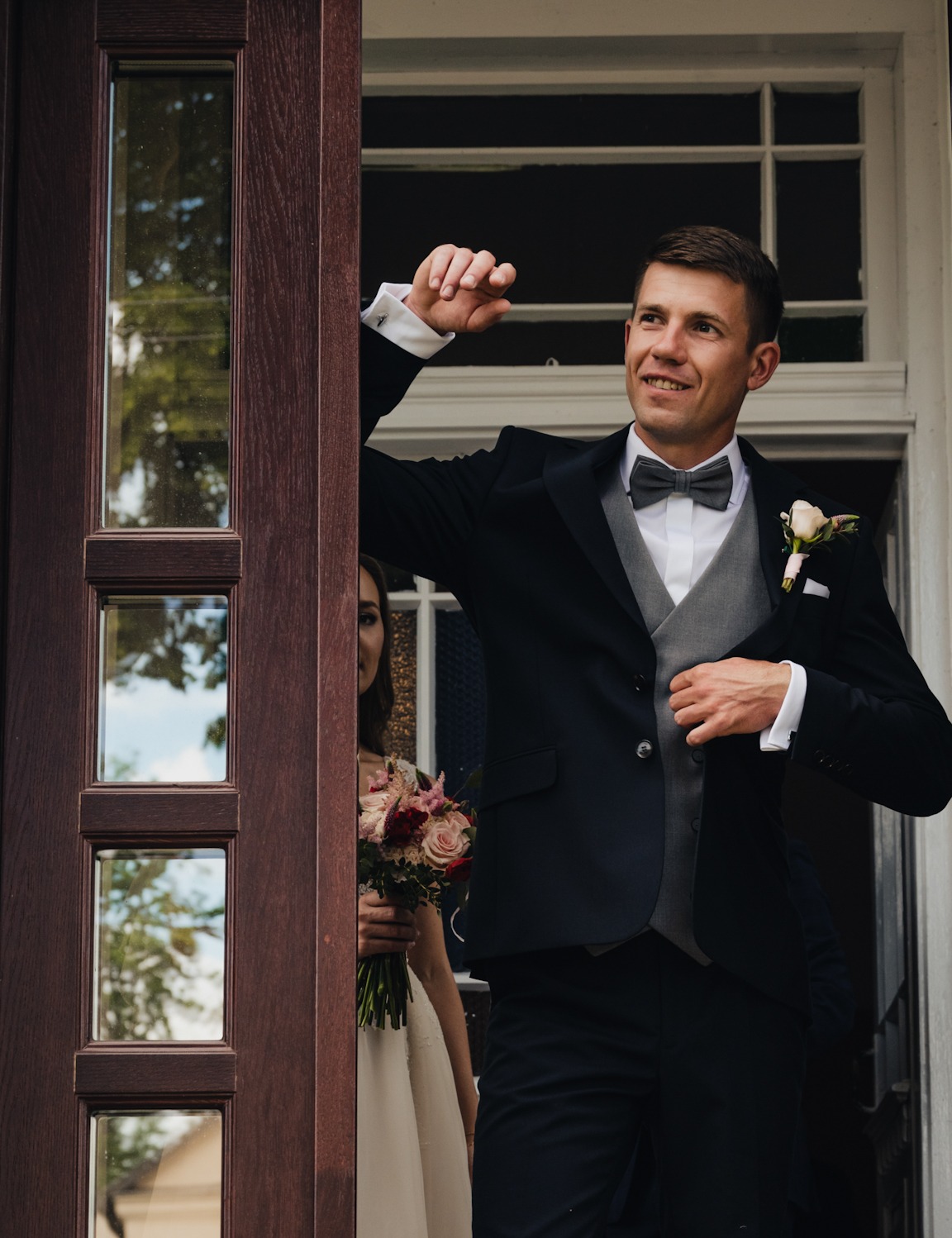 Uśmiechnięty pan młody w ciemnym garniturze i szarej kamizelce, wychodzący z budynku, obok niego w tle widoczna panna młoda z bukietem kwiatów.