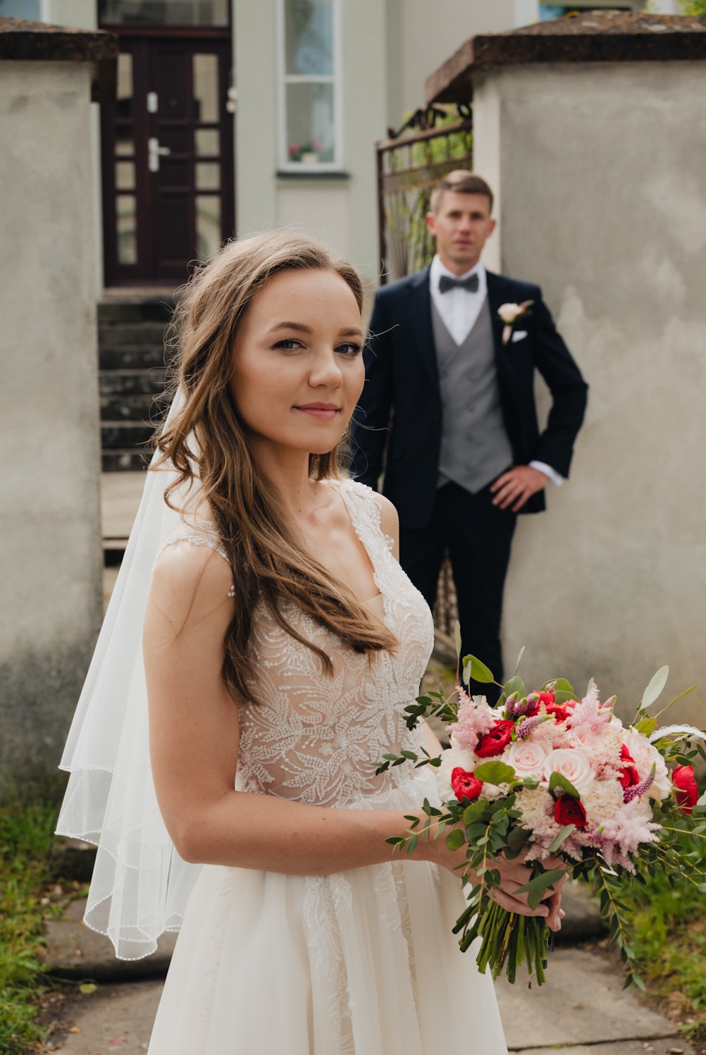 Panna młoda w koronkowej sukni ślubnej z bukietem różowych i czerwonych kwiatów, pan młody w tle w garniturze z muchą.