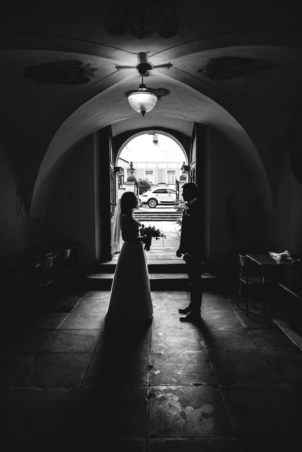Silhouetted bride and groom stand in a dimly lit arched doorway, a white car visible in the background, with a decorative ceiling lamp overhead.