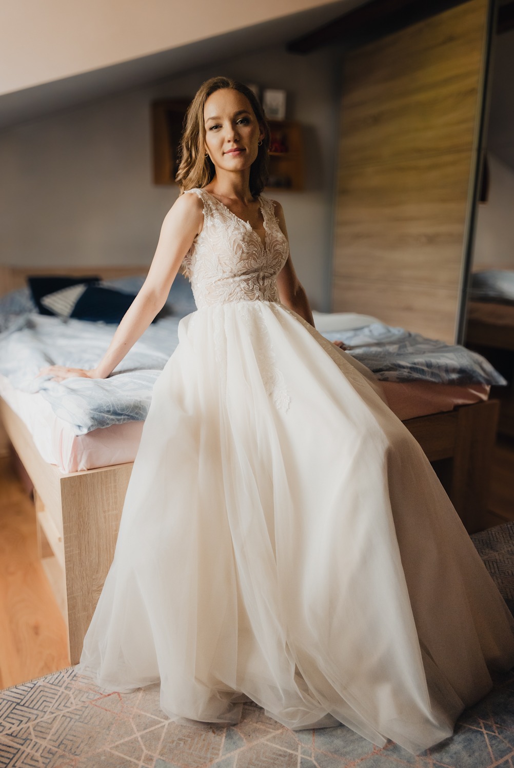 Panna młoda w koronkowej sukni ślubnej pozuje siedząc na łóżku w sypialni, z uśmiechem patrzy w obiektyw.