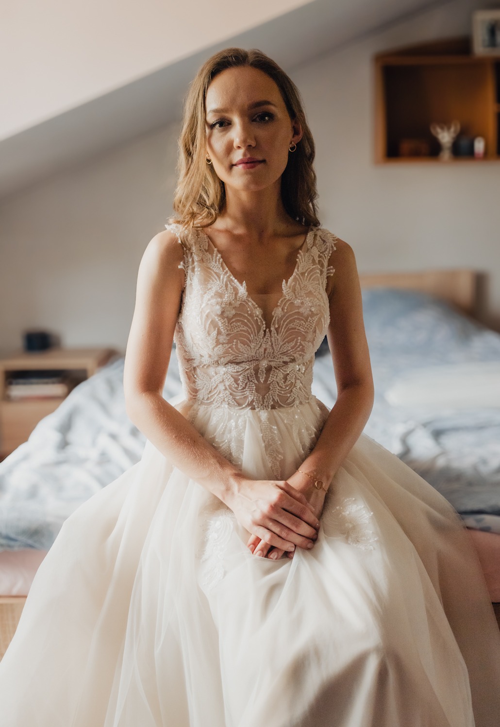 Panna młoda w koronkowej sukni ślubnej siedzi na łóżku w pokoju na poddaszu, z delikatnym uśmiechem i złotą bransoletką na nadgarstku.
