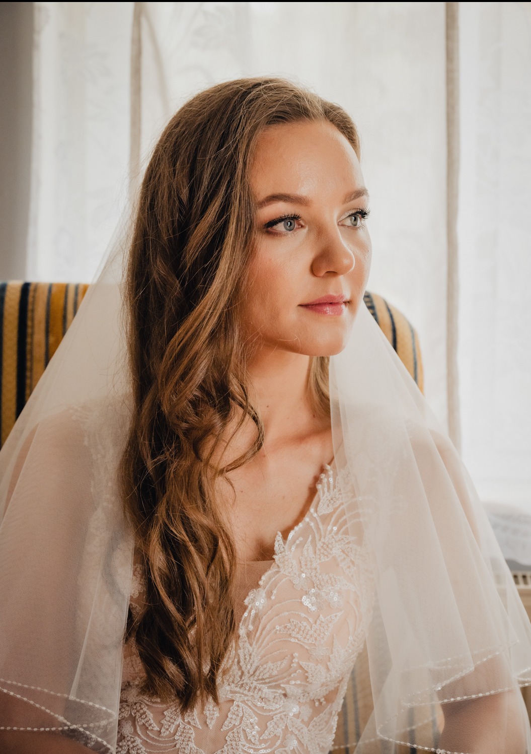 Panna młoda w koronkowej sukni i welonie, siedząca na krześle w paski, zamyślona patrzy w dal przy oknie.