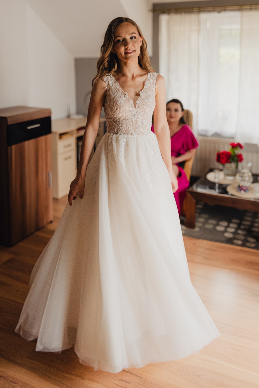 Panna młoda w koronkowej sukni ślubnej z tiulowym dołem, uśmiechnięta, pozuje w jasnym pokoju, w tle siedzi kobieta w różowej sukience.
