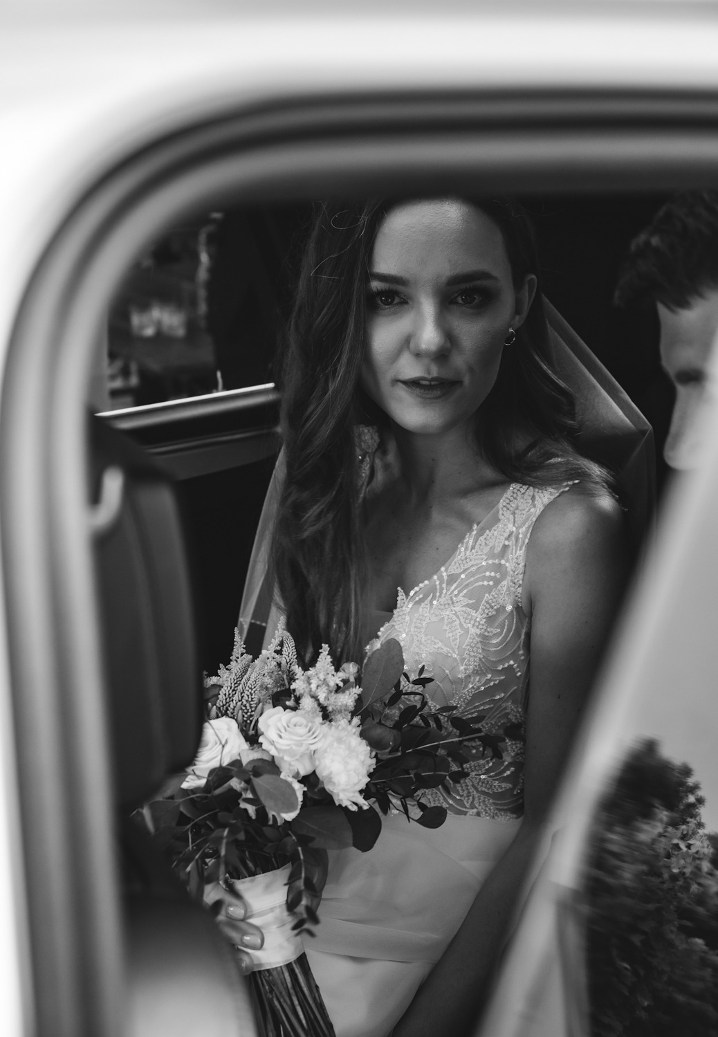 Czarno-białe zdjęcie panny młodej w sukni ślubnej z bukietem, siedzącej w samochodzie i patrzącej w obiektyw.