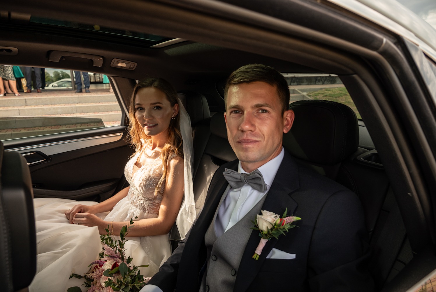 Para młoda w samochodzie z otwartym dachem: panna młoda w sukni z koronkowymi aplikacjami trzyma bukiet, pan młody w ciemnym garniturze z szarą muchą.