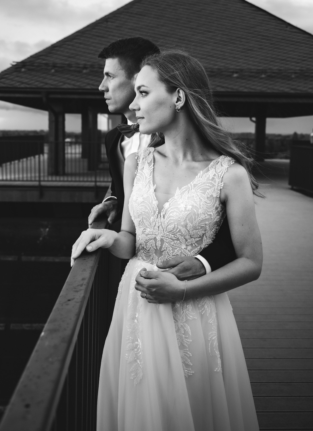 Czarno-białe zdjęcie pary młodej stojącej na drewnianym pomoście, z widokiem na pawilon w tle. Panna młoda w koronkowej sukni, pan młody w smokingu z muchą, oboje patrzą w dal.