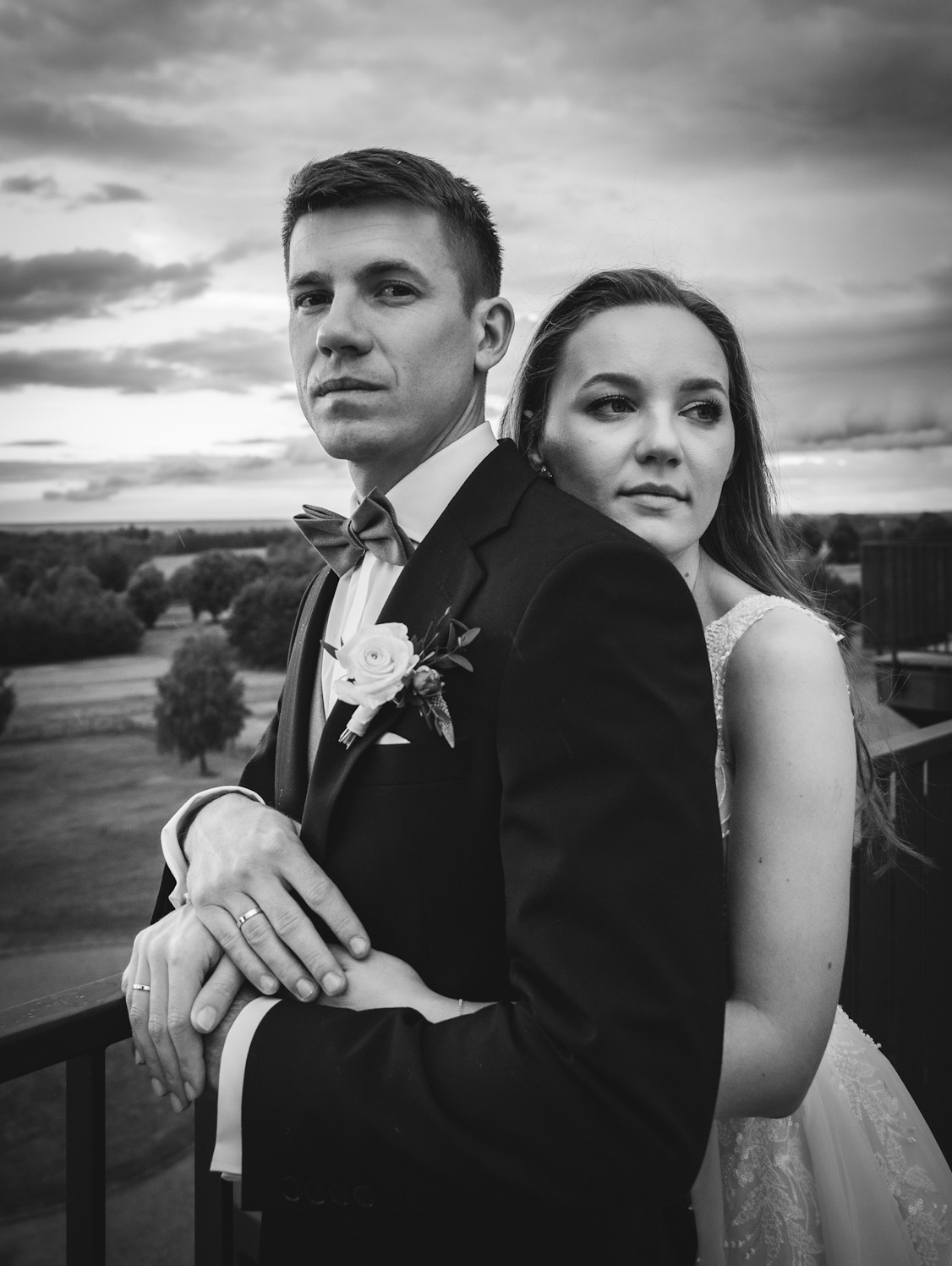 Czarno-białe zdjęcie pary młodej na tle pochmurnego nieba i zieleni. Pan młody w ciemnym garniturze z muszką i butonierką, obejmuje pannę młodą w białej sukni. Widoczne obrączki na palcach.