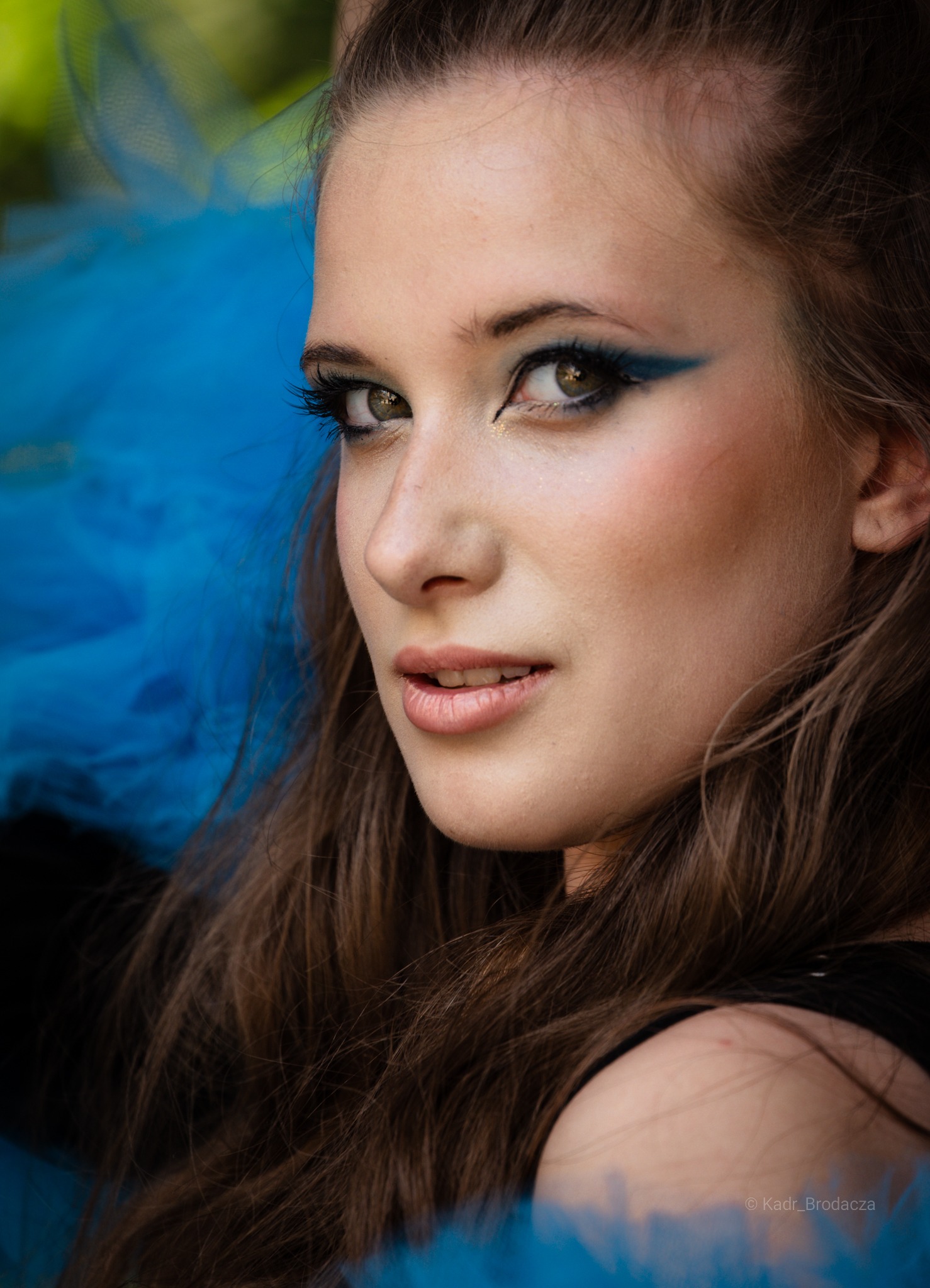 Portret młodej kobiety z intensywnym makijażem oczu w odcieniach błękitu, z błękitnym tiulem w tle, spoglądającej w stronę widza.