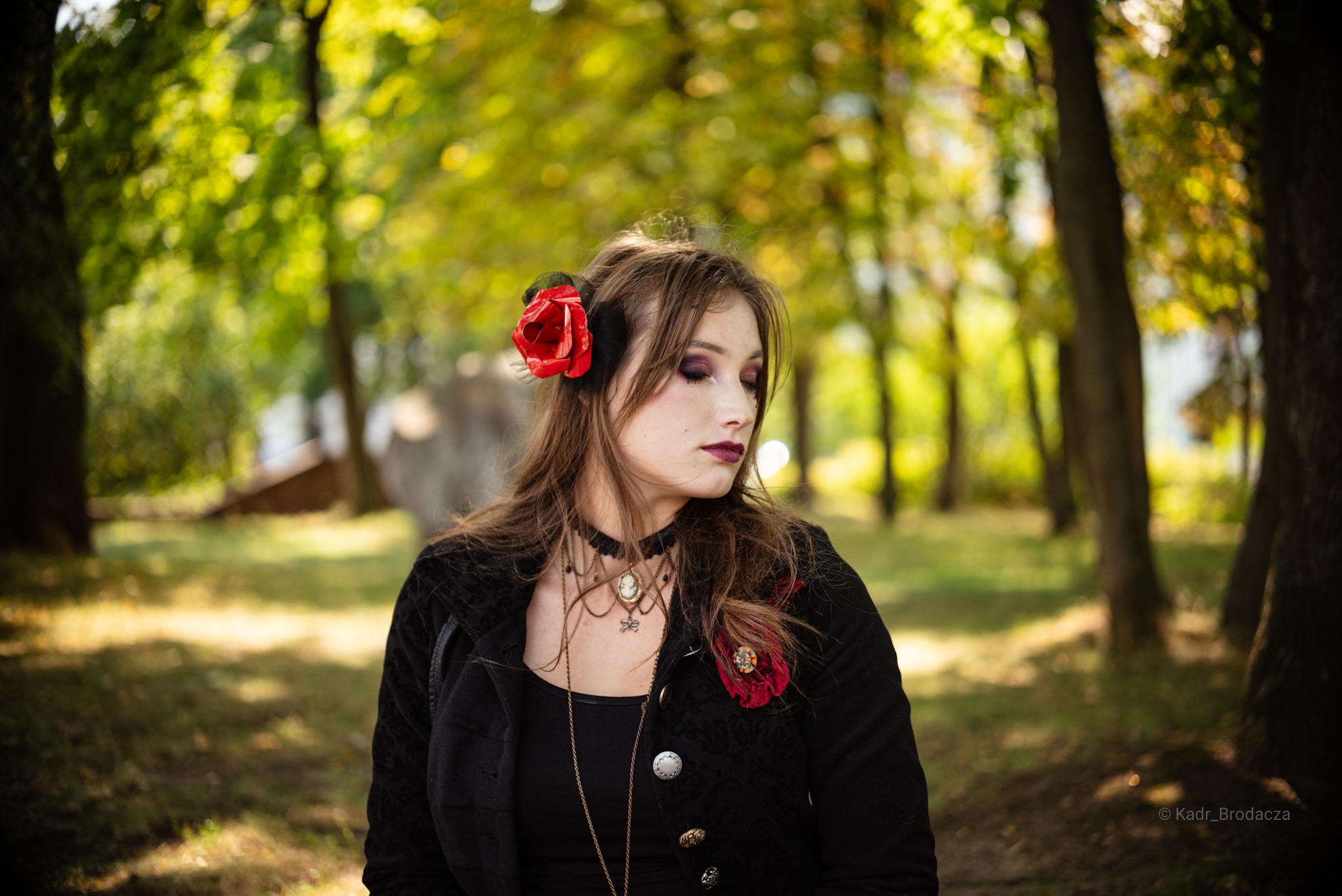 Portret kobiety w czarnej kurtce z różą we włosach, patrzącej w bok na tle drzew w parku. Stylizacja gotycka, makijaż w odcieniach fioletu. Delikatne światło.
