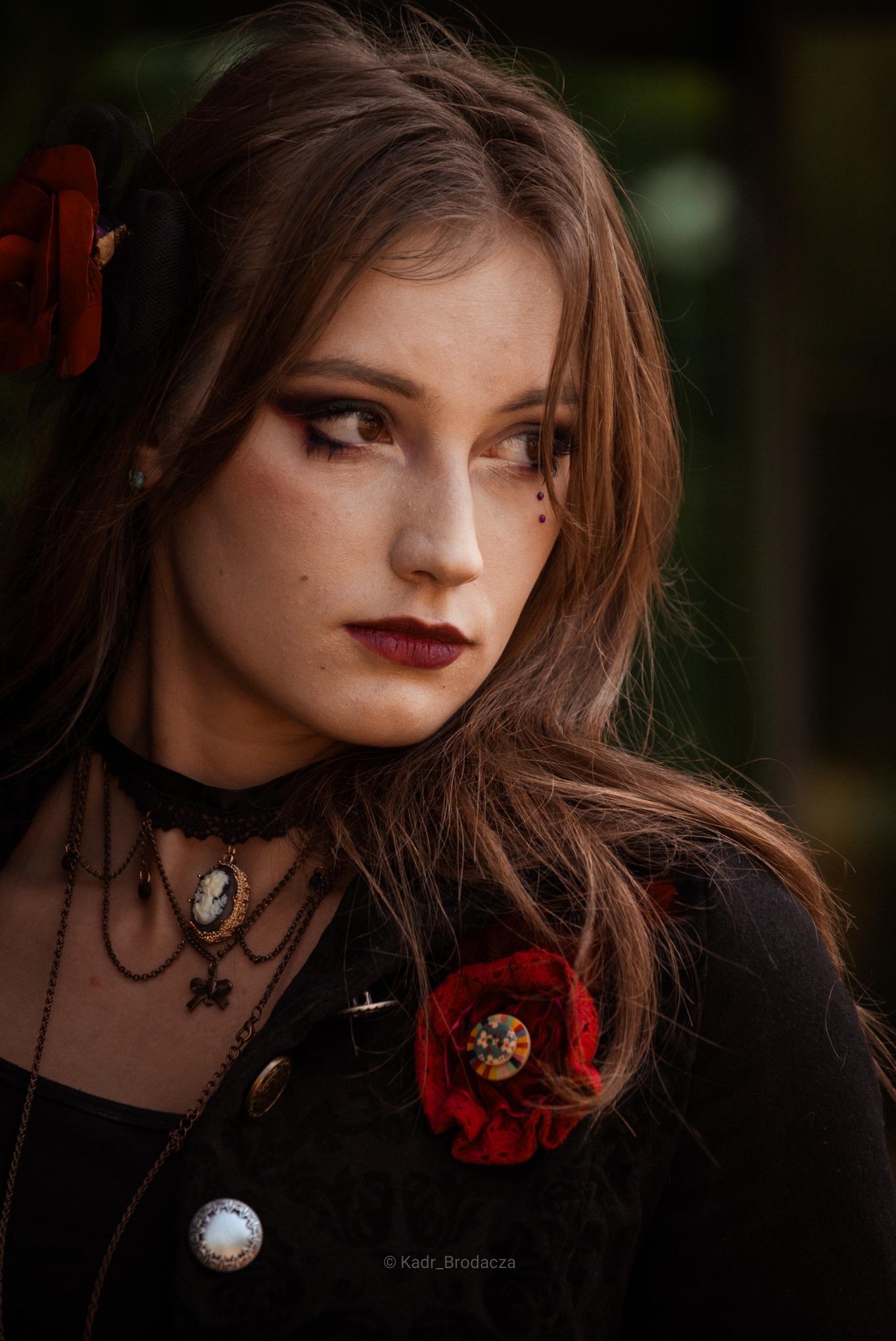 Portret kobiety z artystycznym makijażem i gotycką biżuterią, wpatrującej się w dal, z rozpuszczonymi włosami i kwiatem we włosach.