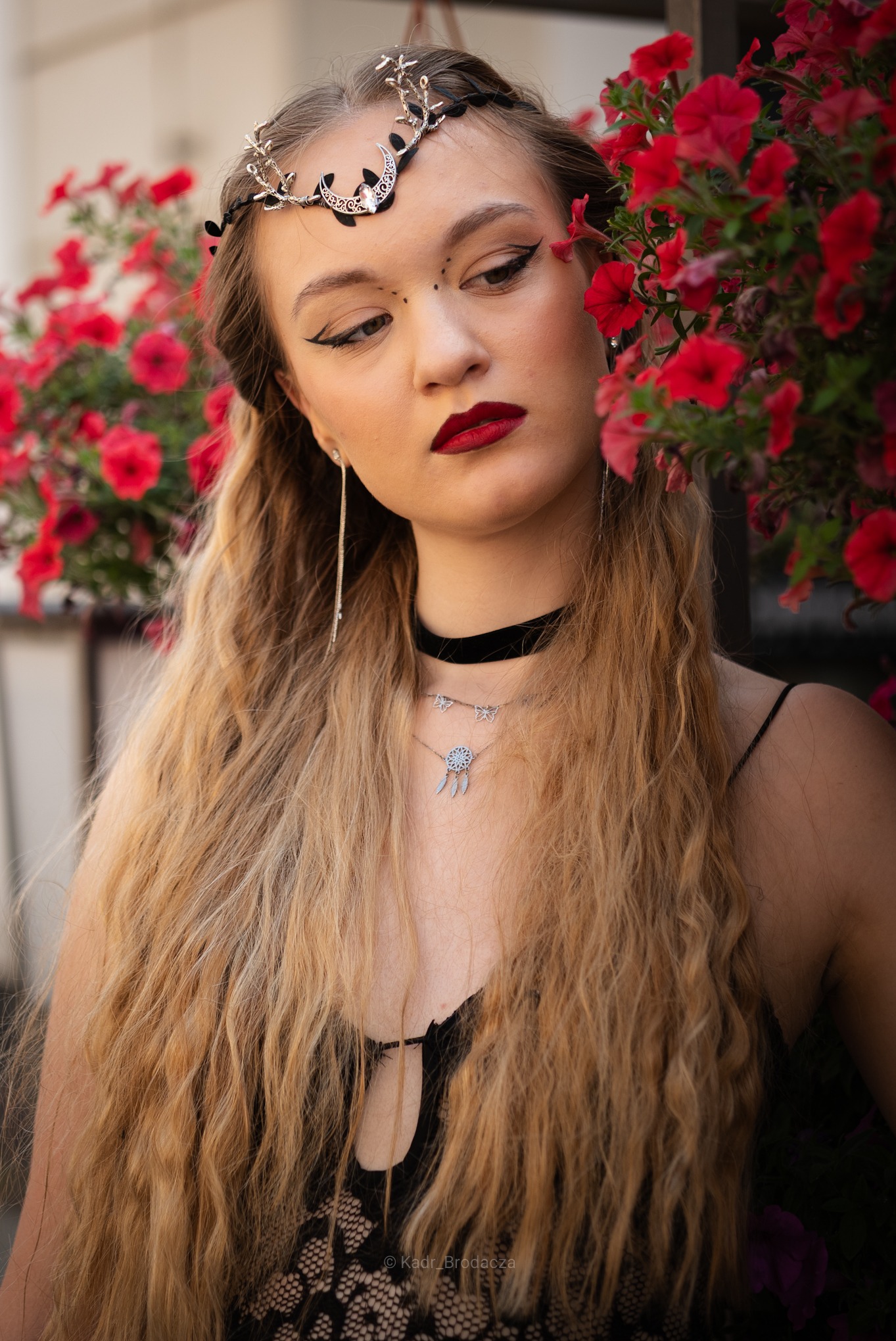 Portret kobiety z długimi blond włosami, w biżuterii i makijażu, pozującej na tle czerwonych kwiatów. Uwagę przyciąga ozdobny diadem na głowie i wyrazisty makijaż.