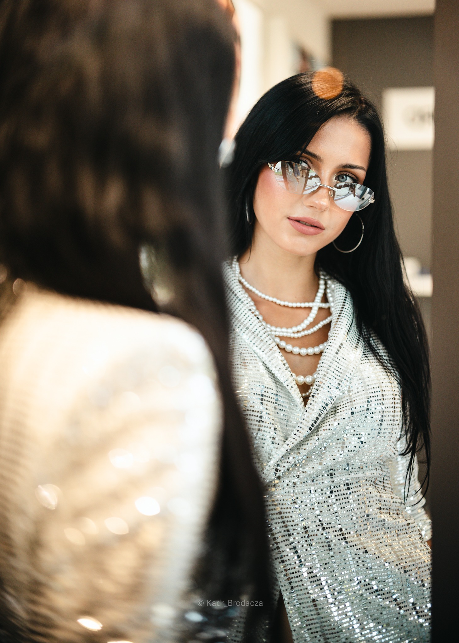 Kobieta z czarnymi włosami w srebrnej, cekinowej marynarce i okularach w kształcie serc, pozuje przed lustrem, prezentując stylizację na sesję kobiecą.