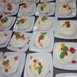 Rząd białych bezik&oacute;w na kwadratowych talerzykach, dekorowanych bitą śmietaną, kostkami owoc&oacute;w i listkiem mięty, widok z g&oacute;ry.