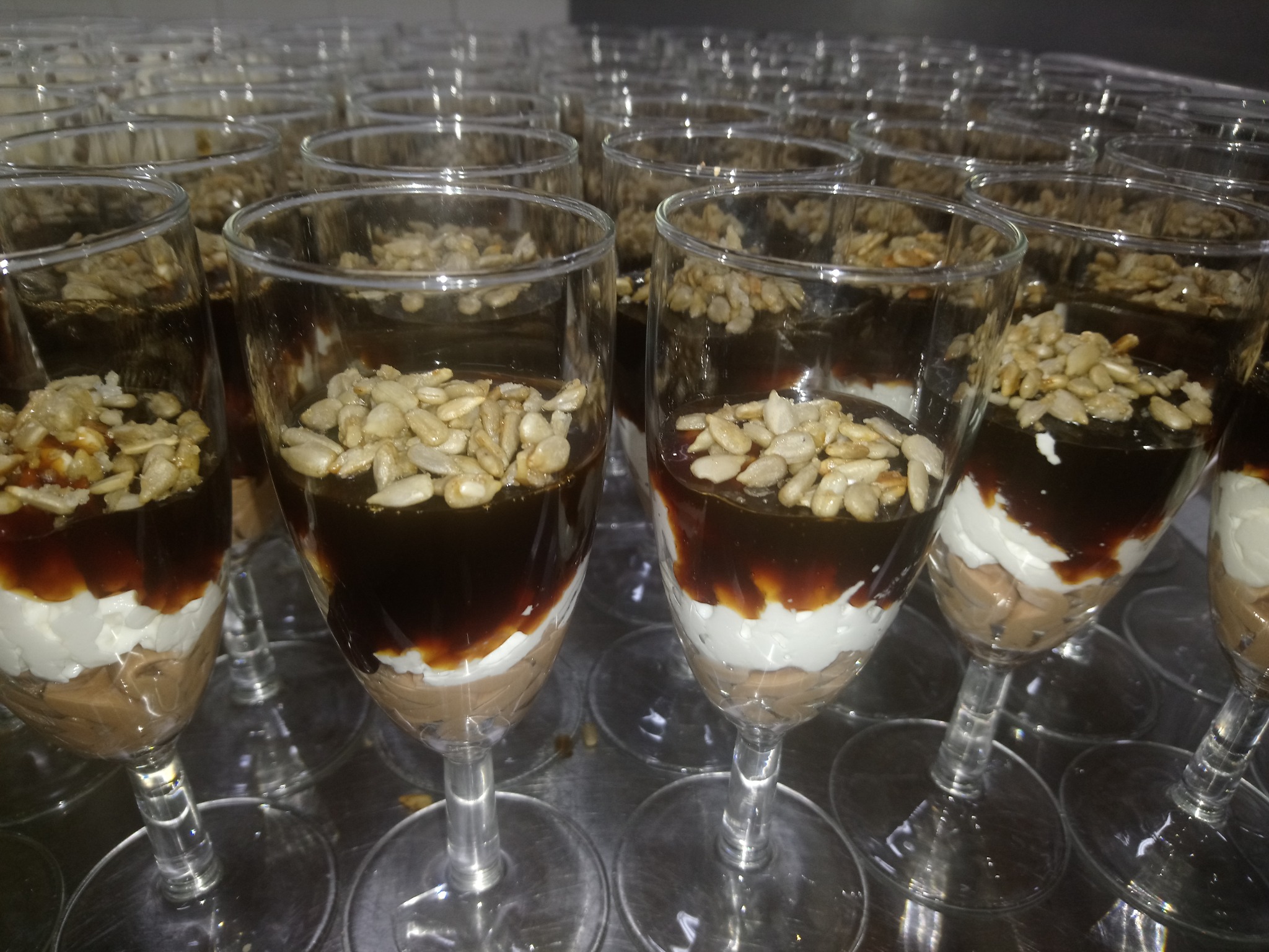 Deser w kieliszku: warstwy kremu czekoladowego, bitej śmietany, polewy karmelowej i posypki z ziaren słonecznika, ustawione w rzędach na metalowej tacy.