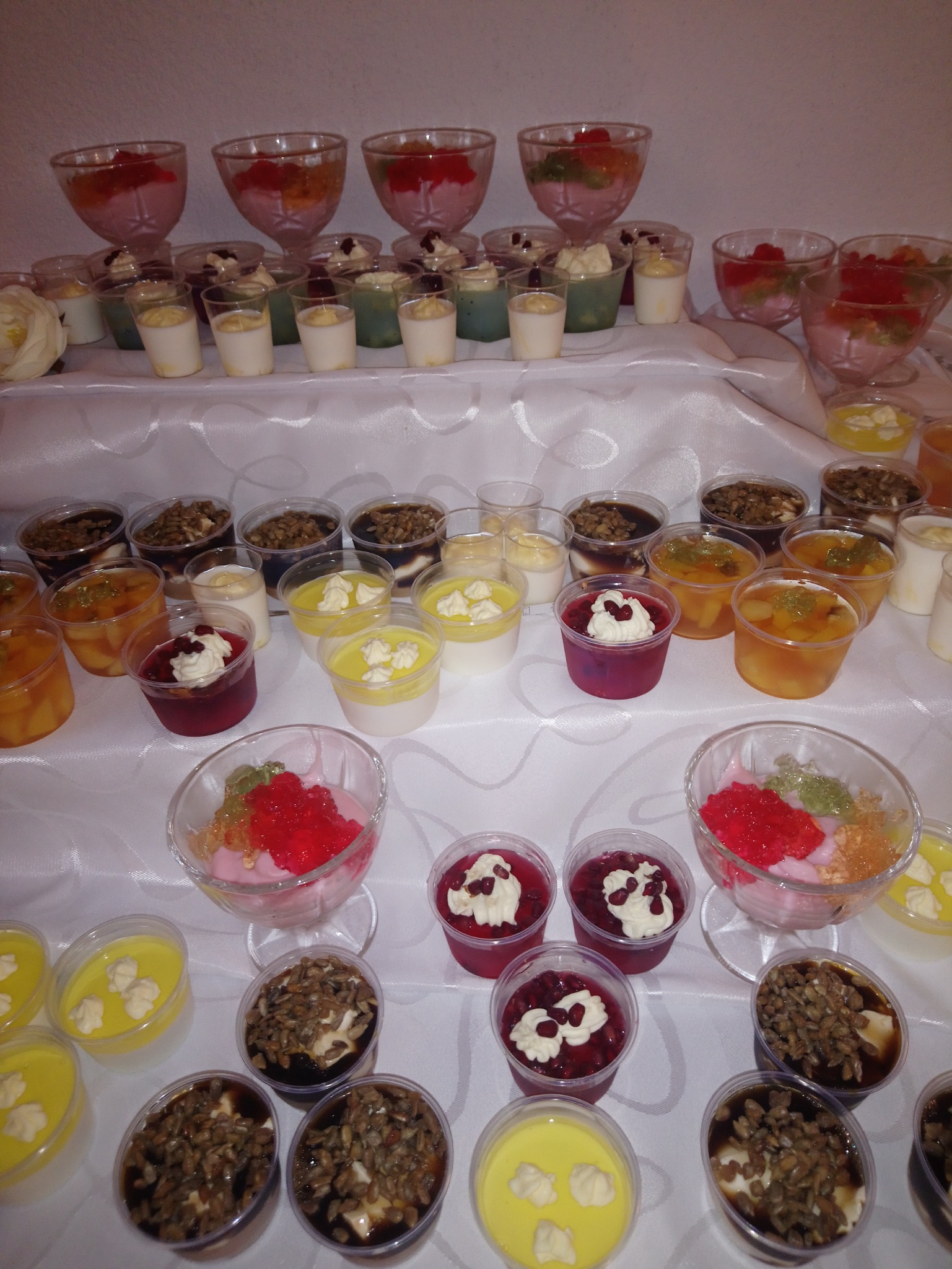 Słodki bufet z różnorodnymi deserami w szklanych i plastikowych naczyniach, na białym obrusie.