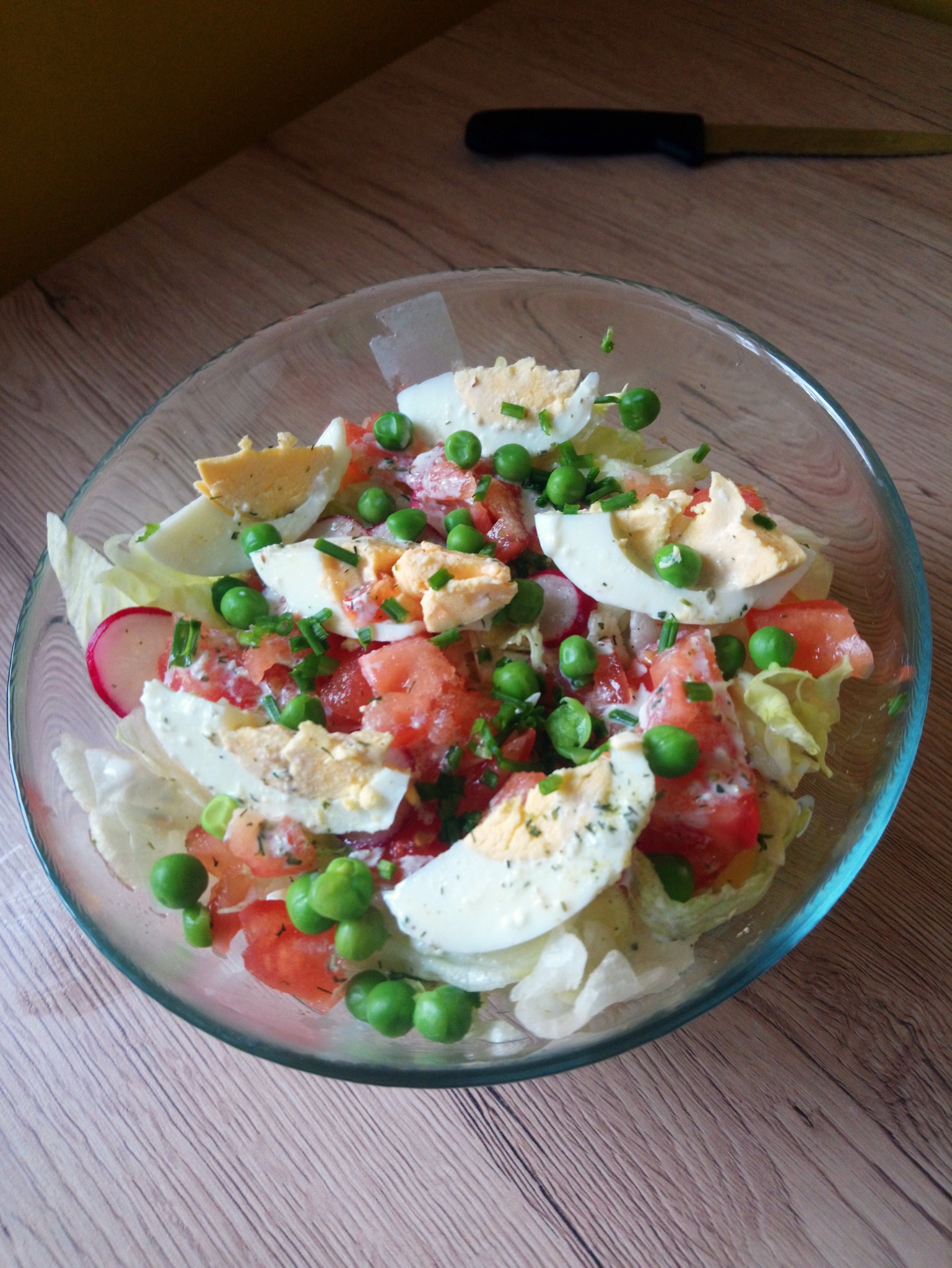 Szklana misa z sałatką warzywną: pomidory, jajka na twardo, groszek, rzodkiewka i sałata lodowa, całość posypana szczypiorkiem, w tle nóż na drewnianym blacie.