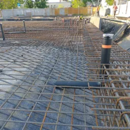 Zbrojenie fundamentów z widocznymi rurami instalacyjnymi i pracownikami w tle.