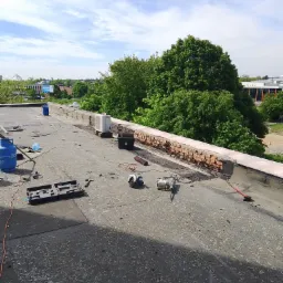 Prace dekarskie na płaskim dachu: uszkodzona obróbka blacharska, narzędzia dekarskie (szlifierka, młot udarowy), butle gazowe, widok na zieleń miejską.