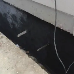 Fundament budynku po izolacji bitumicznej, widoczne wystające pręty zbrojeniowe i fragment ściany zewnętrznej.