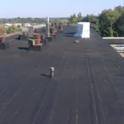 Czarny, płaski dach pokryty papą termozgrzewalną z widocznymi kominkami wentylacyjnymi i zabezpieczonym pakunkiem materiałów budowlanych, w tle zieleń drzew i błękitne niebo.