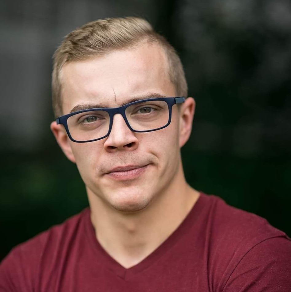 Portret młodego mężczyzny w okularach z niebieskimi oprawkami i bordowej koszulce, lekki uśmiech, ciemne tło.