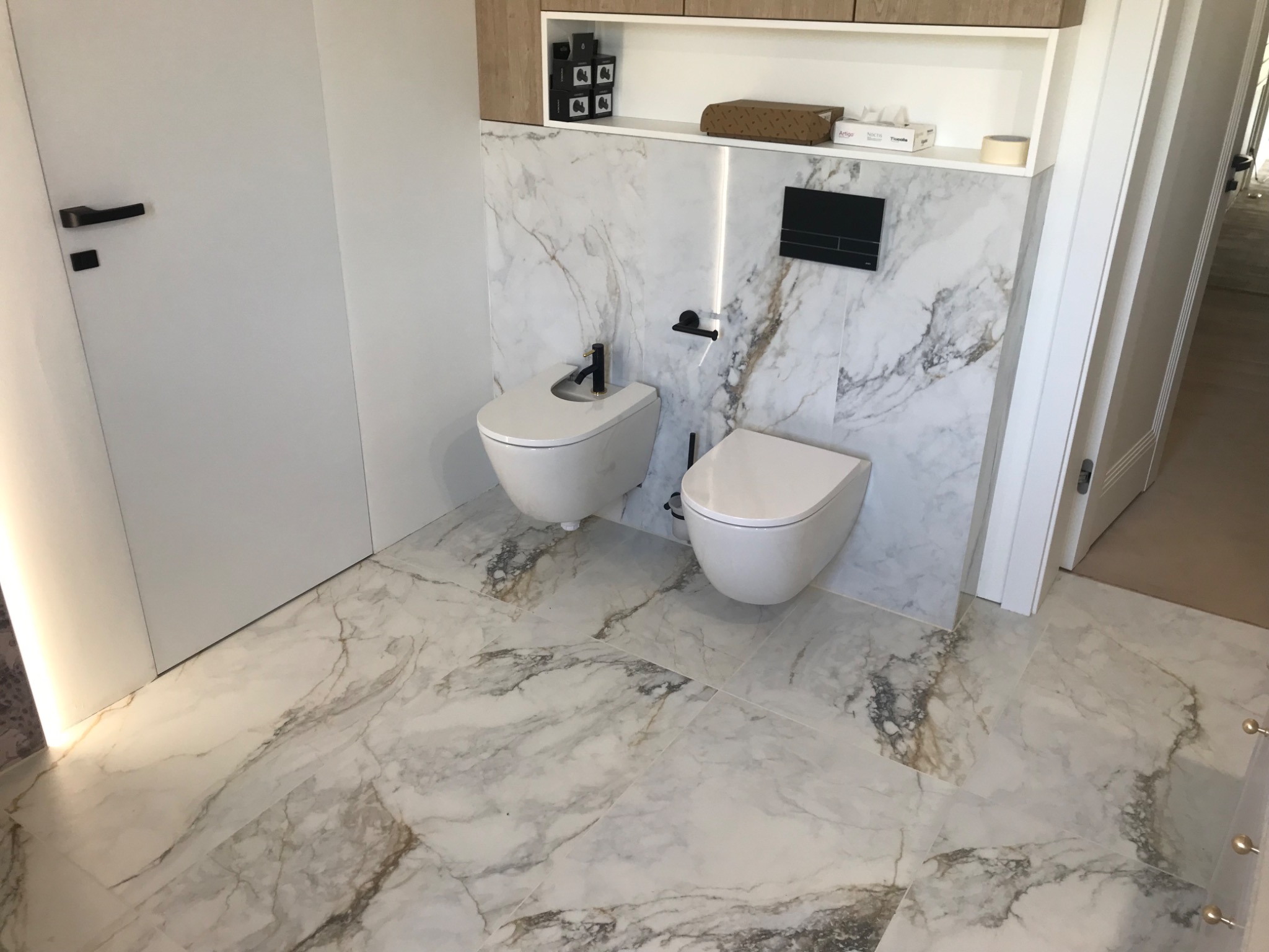 Nowoczesna łazienka z marmurowymi płytkami na ścianach i podłodze, wisząca toaleta i bidet z czarną armaturą, białe drzwi z czarną klamką i zabudowana szafka z półkami.