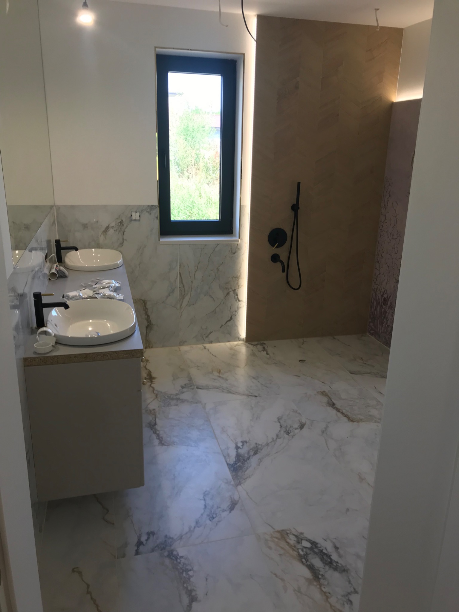 Łazienka w trakcie wykańczania: marmurowe płytki na ścianach i podłodze, dwie białe umywalki z czarnymi bateriami, widoczne akcesoria montażowe, kabina prysznicowa z drewnopodobnymi płytkami...